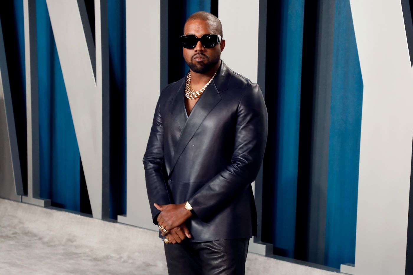Kanye West wearing a black leather suit jacket and pants with sunglasses in front of a white and blue background.