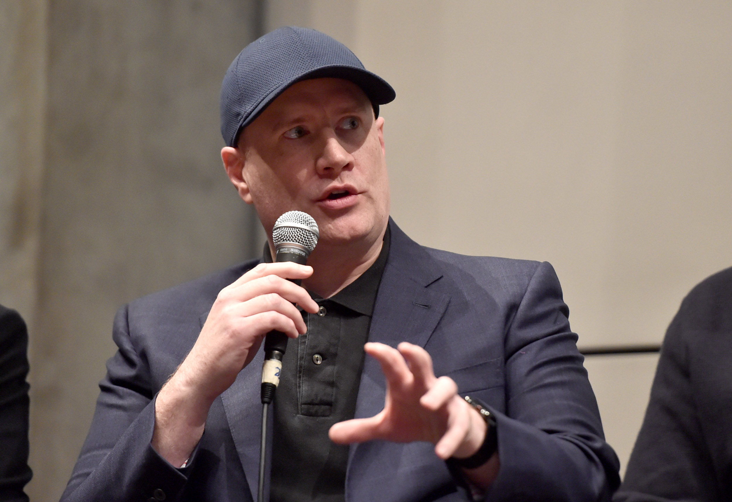 Marvel Studios president Kevin Feige wearing a grey suit and hat and talking into a microphone