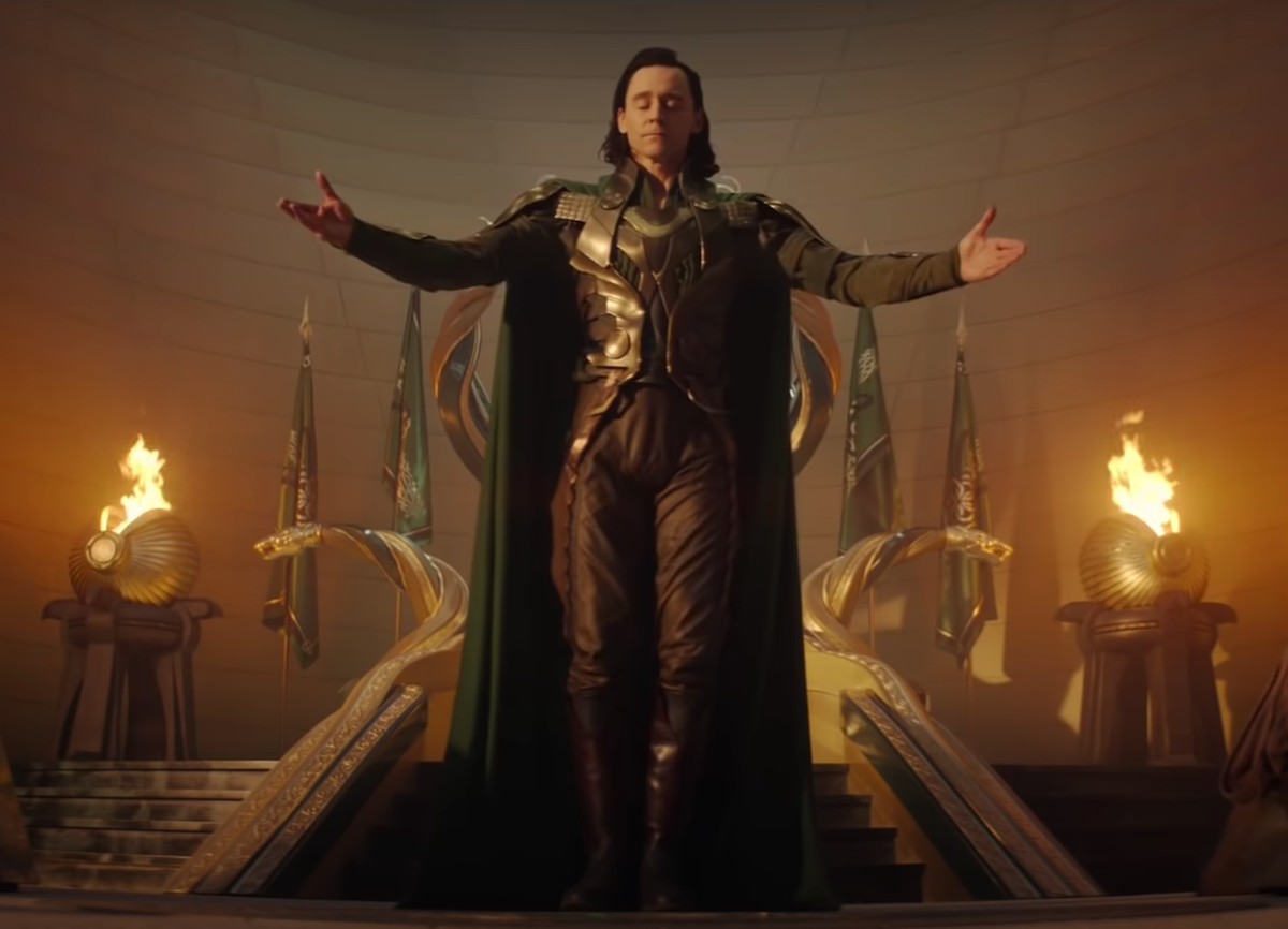 Tom Hiddleston in 'Loki.' He stands in front of a gold throne in what resembles Asgard. He wears a regal green cape and an armored green, gold, and black suit. His arms are outstretched as if accepting praise and his eyes are closed.