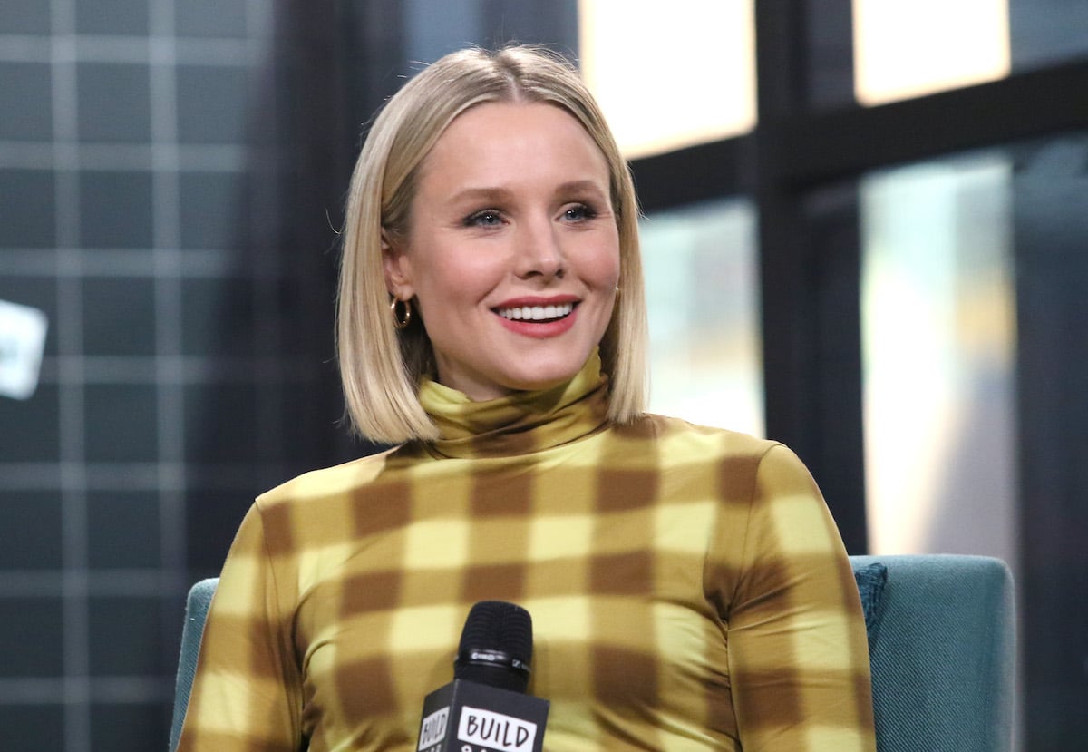 Kristen Bell sits on stage is a yellow and brown checkered outfit. She holds a microphone.