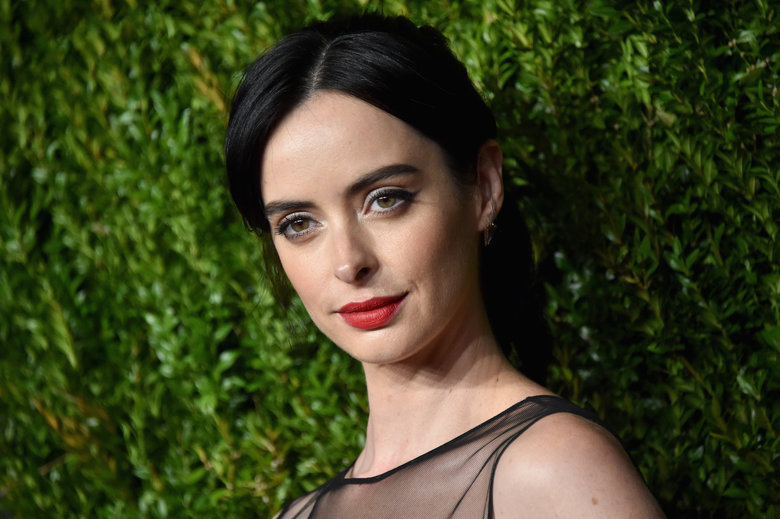 Krysten Ritter stands in front of greenery wearing a black sheer dress and red lipstick