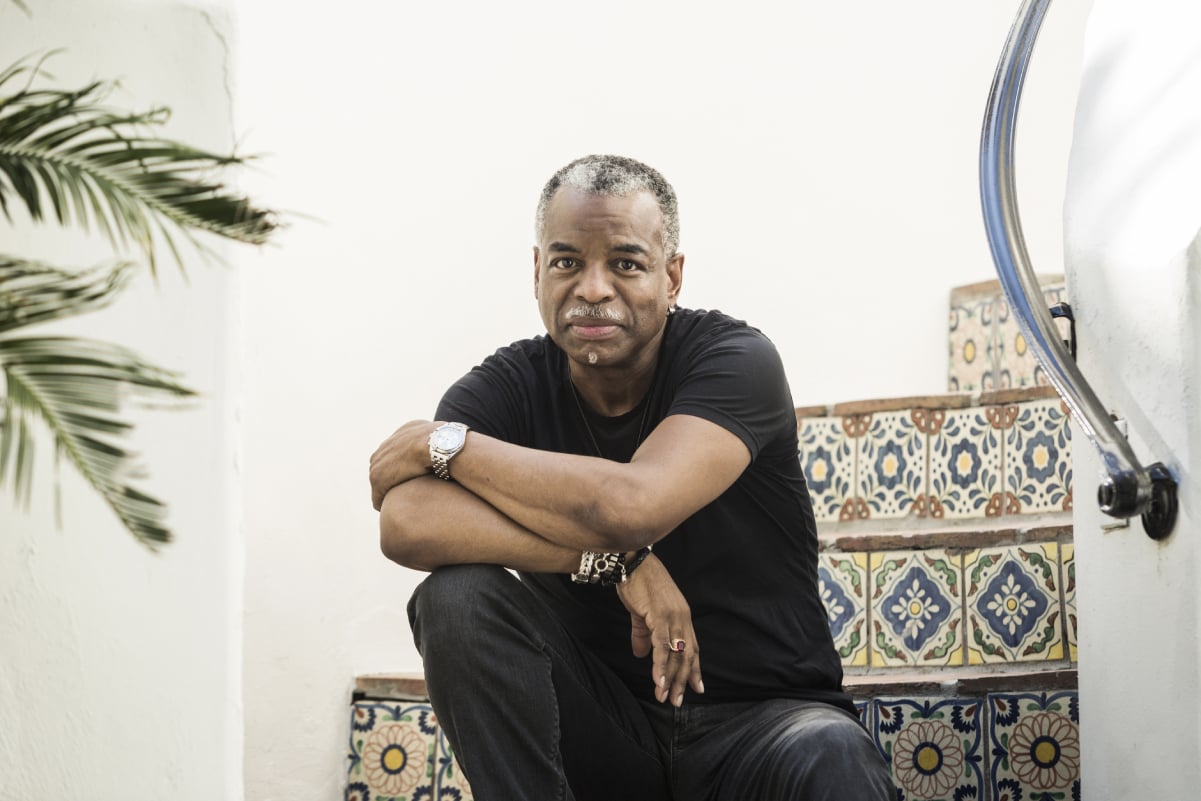 Actor LeVar Burton sits for a portrait in black pants and shirt with his arm crossed over his knee.