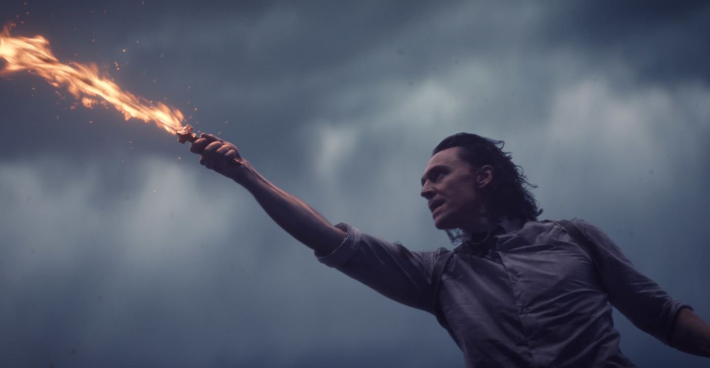 Loki wearing a white button up shirt with the sleeves rolled up with a flaming sword in his hand lunging to the side.