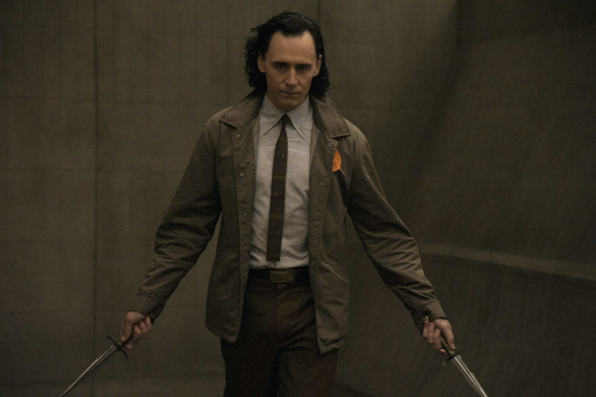 Tom Hiddleston holding a dagger in each hand in 'Loki' on Disney+. He wears a shirt and tie with a brown coat while walking down a brown hallway.