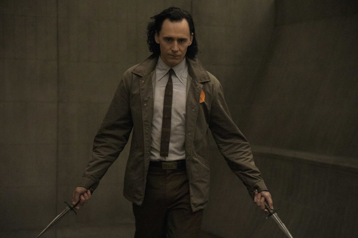 Tom Hiddleston holding a dagger in each hand in 'Loki' on Disney+. He wears a shirt and tie with a brown coat while walking down a brown hallway.