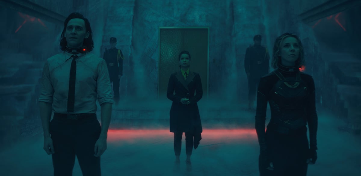 Tom Hiddleston, Sophia Di Martino, and Gugu Mbatha-Raw in 'Loki' Episode 4. Hiddleston wears a shirt and tie and Di Martino wears a black and green armored body suit. Both have electronic bands around their necks. Mbatha-Raw wears a brown and orange TVA uniform. They stand in a smoky room looking up at the Time-Keepers, off-camera.