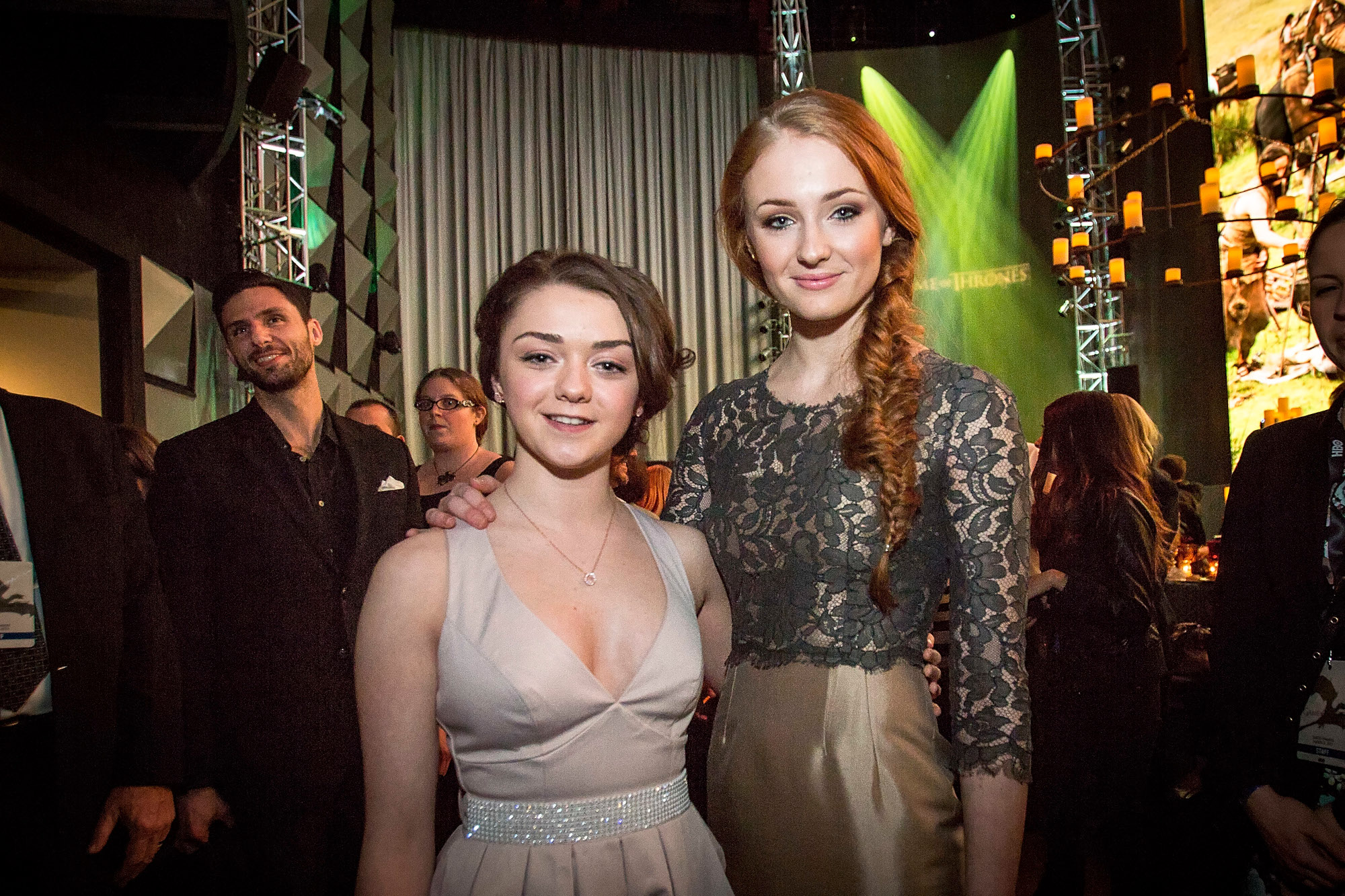 Game of Thrones stars Maisie Williams and Sophie Turner stand side by side. Williams wears a beige dress and Turner wears a lacy top and gold skirt