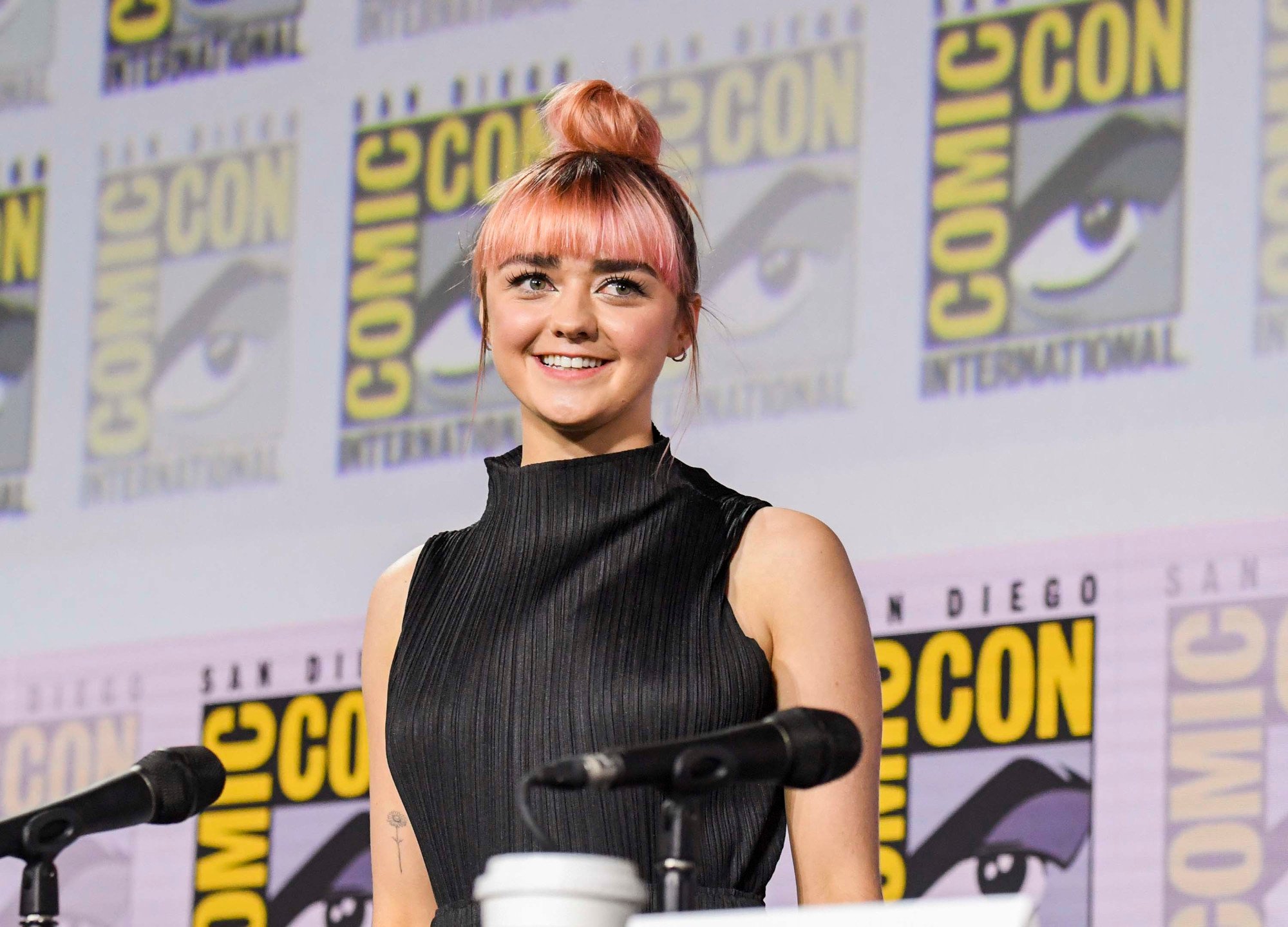 Game of Thrones star Maisie Williams stands in front of a Comic-Con wall with her pink hair in a bun and a black blouse on