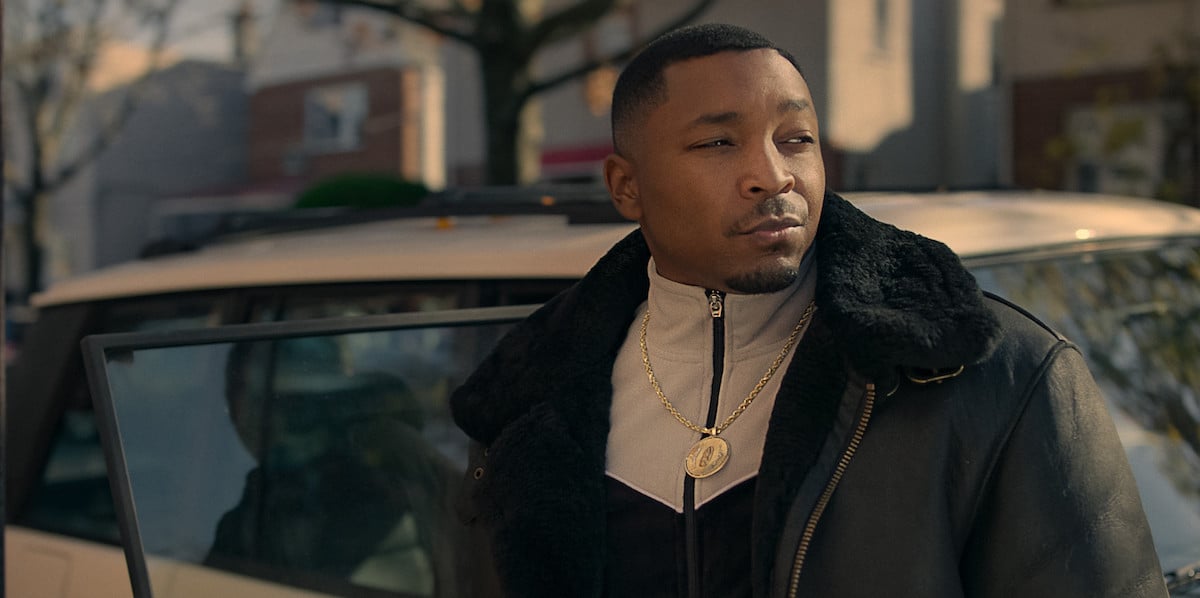 Malcolm Mays as Lou Lou standing outside of a car wearing a coat and chain in 'Power Book III: Raising Kanan'