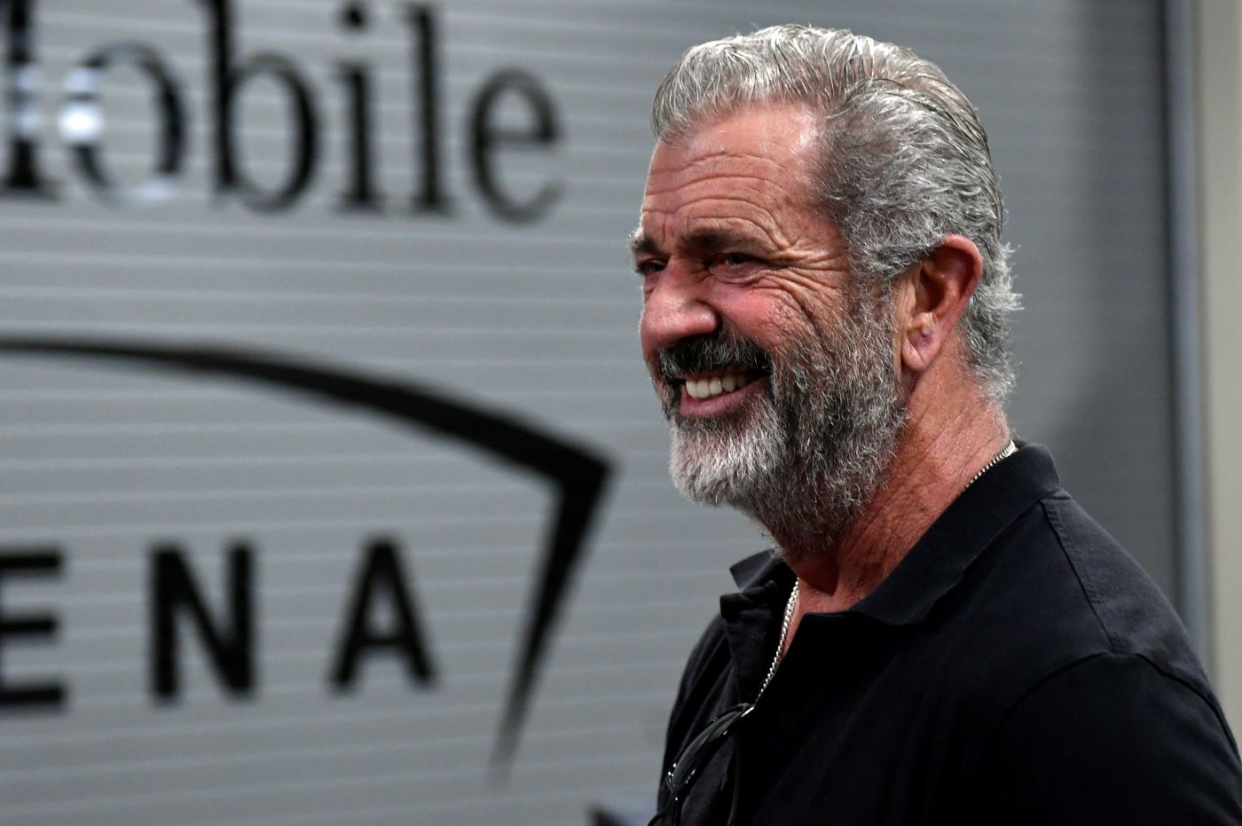 Mel Gibson stands in front of a rippled tin wall wearing a black polo.