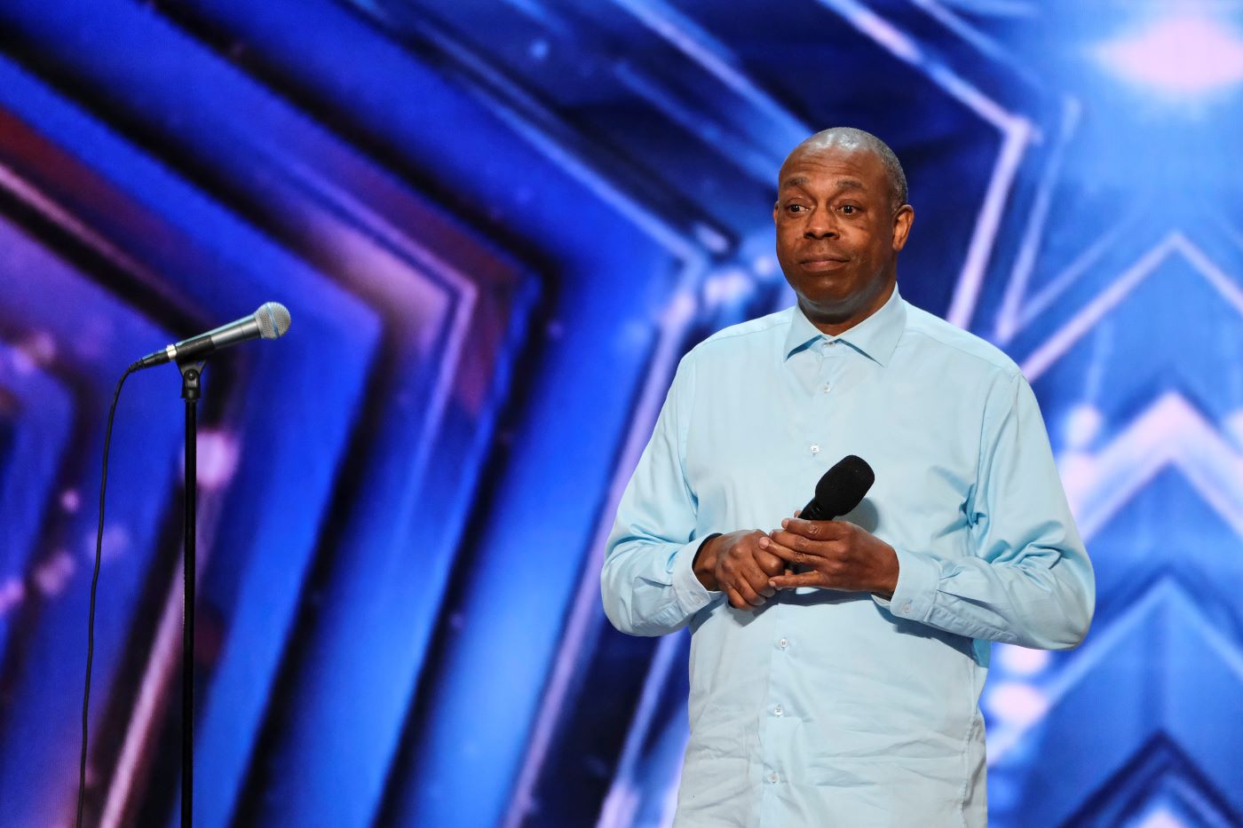 Michael Winslow is a sky blue button-up shirt holding a black microphone in front of a blue and purple textured background.