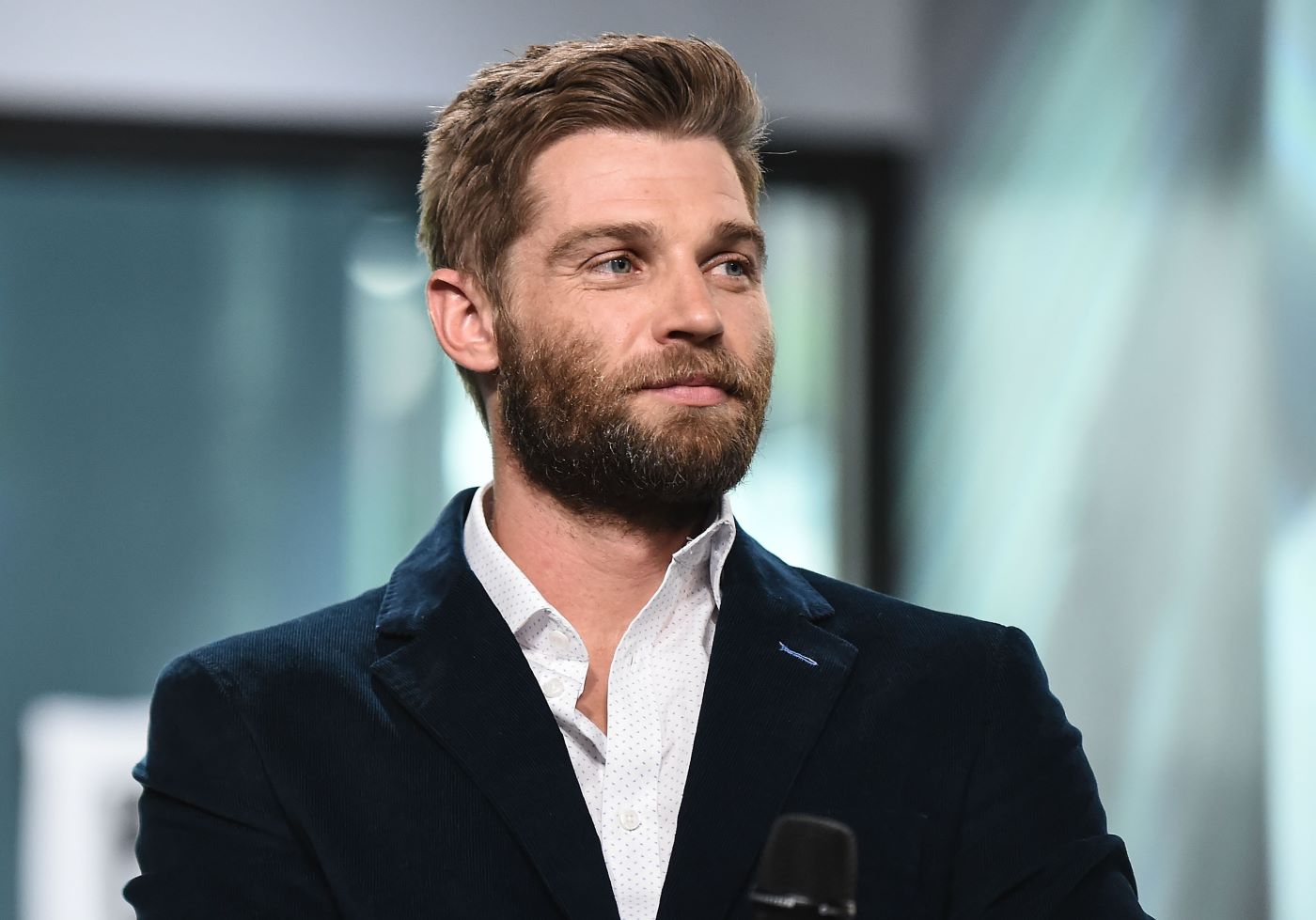 Mike Vogel dressed in a white shirt and a black jacket in front of a very blurred back ground of what looks like a window.