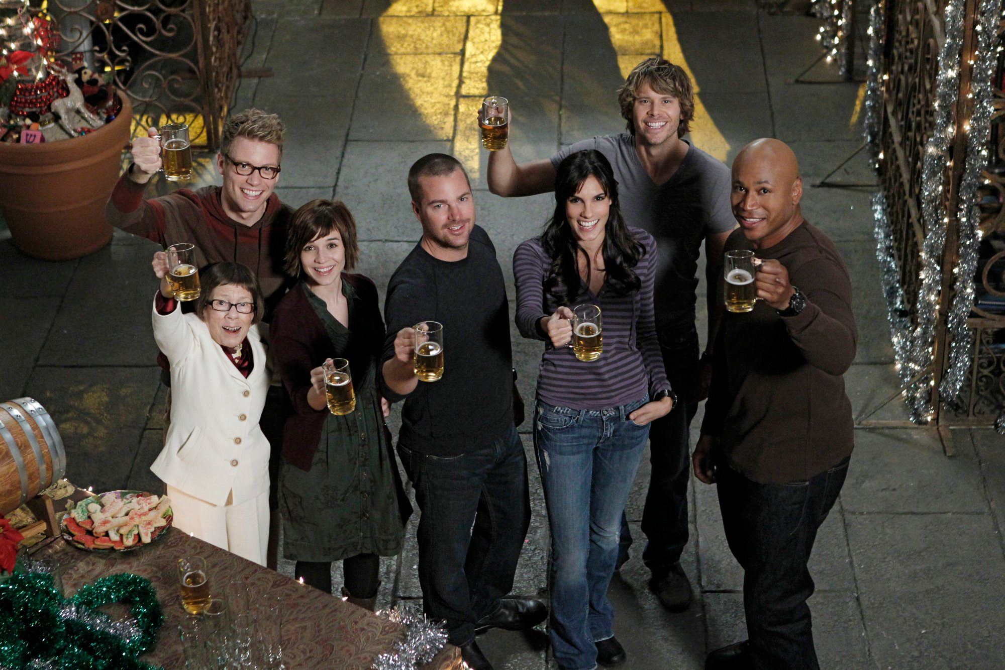 The NCIS Los Angeles cast raises glasses of beer while standing inside the NCIS LA set. 