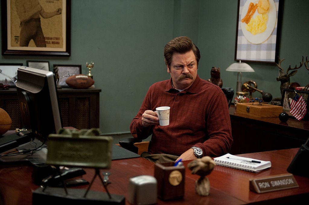 Nick Offerman as Ron Swanson drinks his coffee from behind his desk with a scowl on his face.