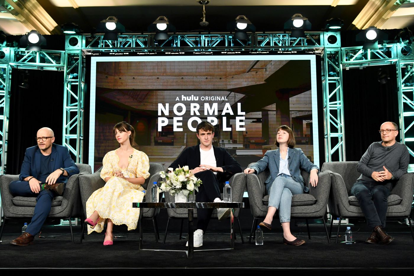 Five cast members sit on a stage in gray chairs behind a black table with water bottles and white flowers in a white vase. The background has industrial beams forming a frame around a projection of an indoor scene with the text, 'A hulu original Normal People' placed on the screen.