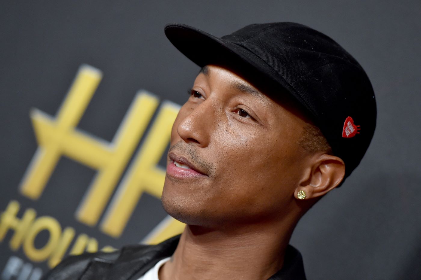 Pharrell Williams in a black ball cap and black jacket with a white undershirt standing in front of a black background with yellow writing.