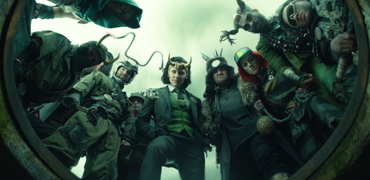 Tom Hiddleston as President Loki surrounded by other Lokis in 'Loki' Episode 5. The group looks down into the opening of an underground tunnel.