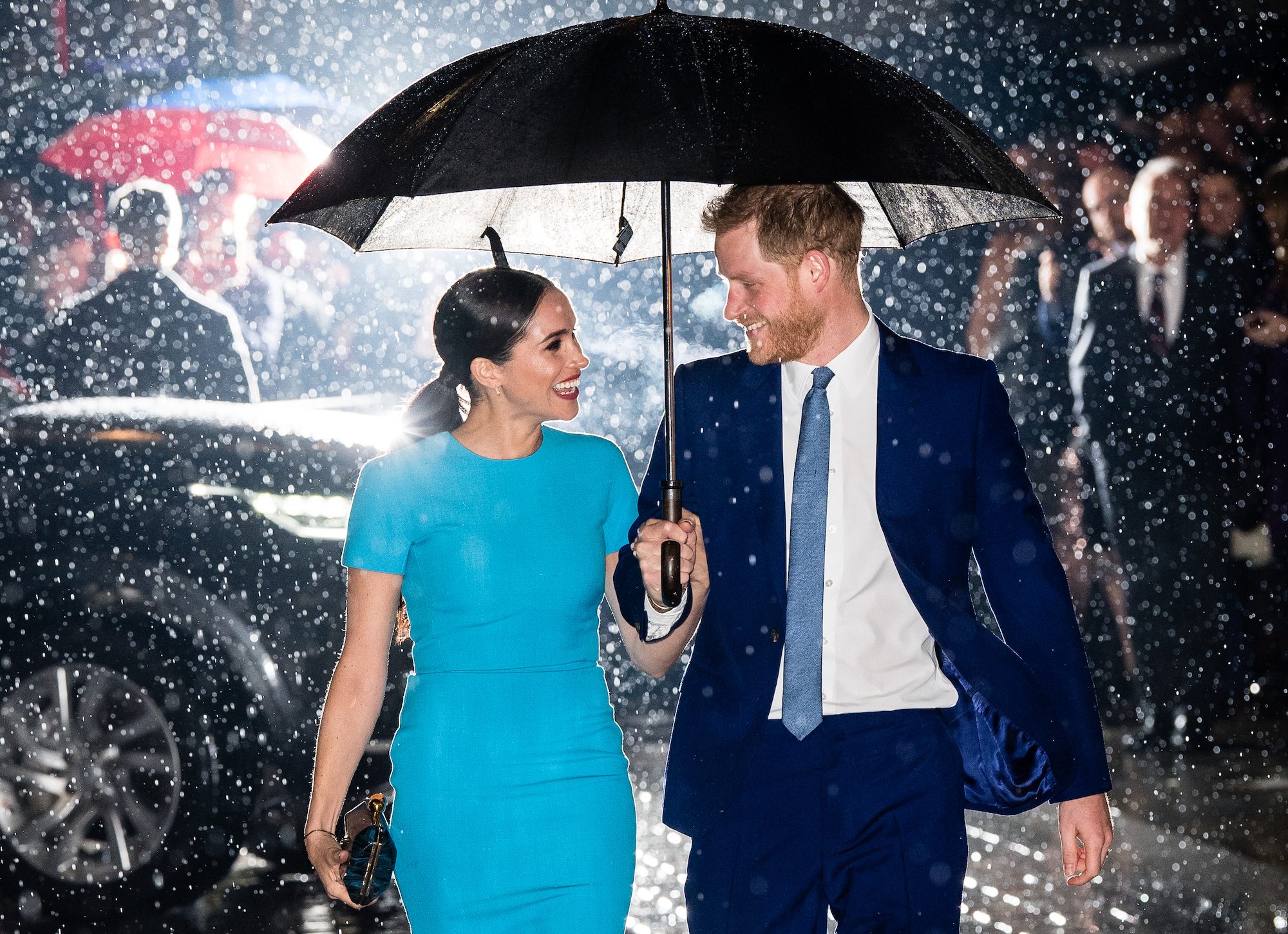 Harry and Meghan smiling at each other under and umbrella, walking in the rain