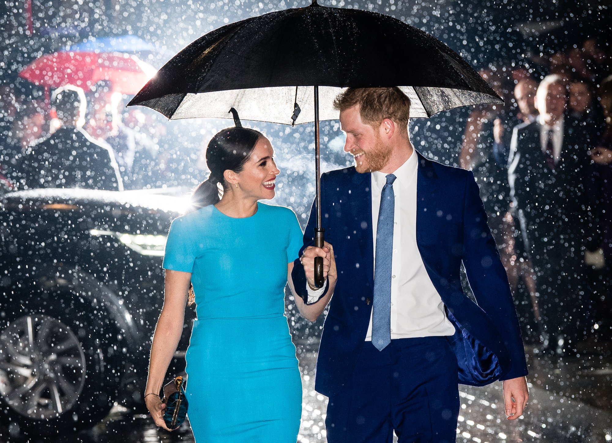 Prince Harry and Meghan Markle smiling at each other under and umbrella, walking in the rain