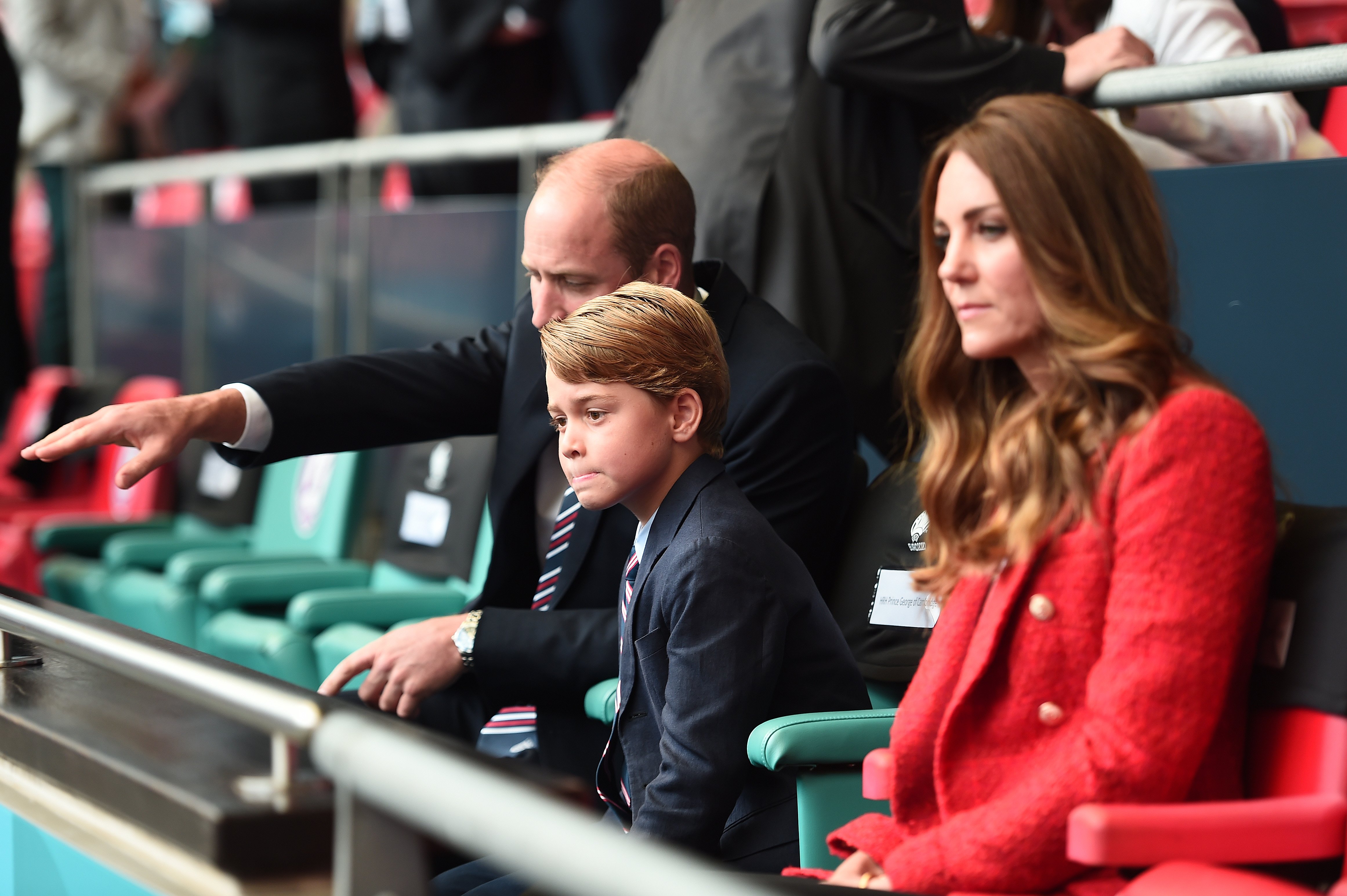 Prince William explaining something to Prince George as they sit in the stands with Kate Middleton at a football match