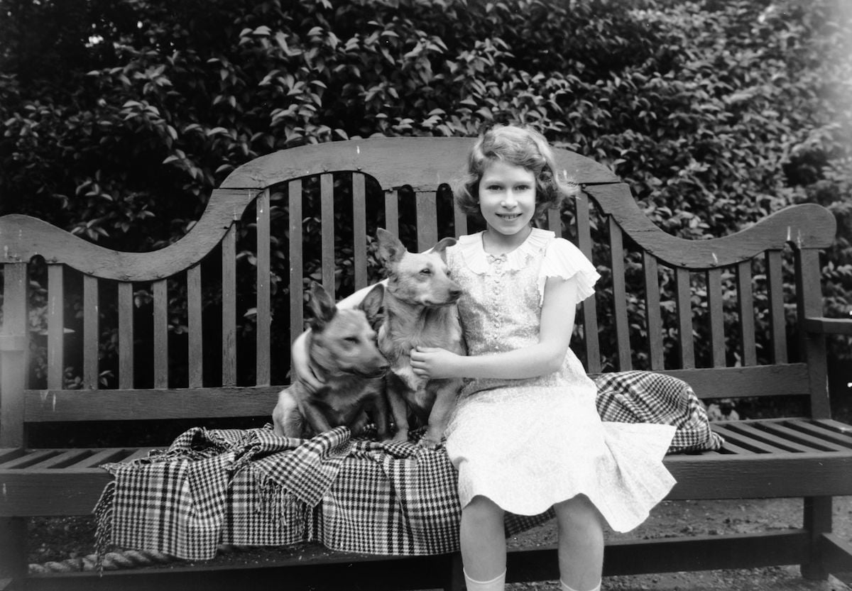 Queen Elizabeth as child, then Princess Elizabeth, sitting on a garden seat with two corgi dogs at her home on 145 Piccadilly, London.