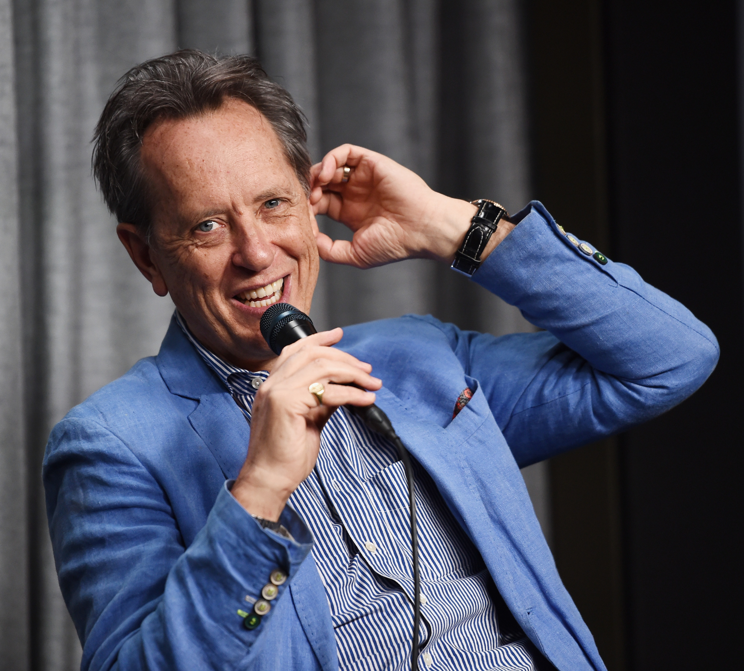 Richard E. Grant wearing a blue shirt, raising one arm, and holding a microphone in the other