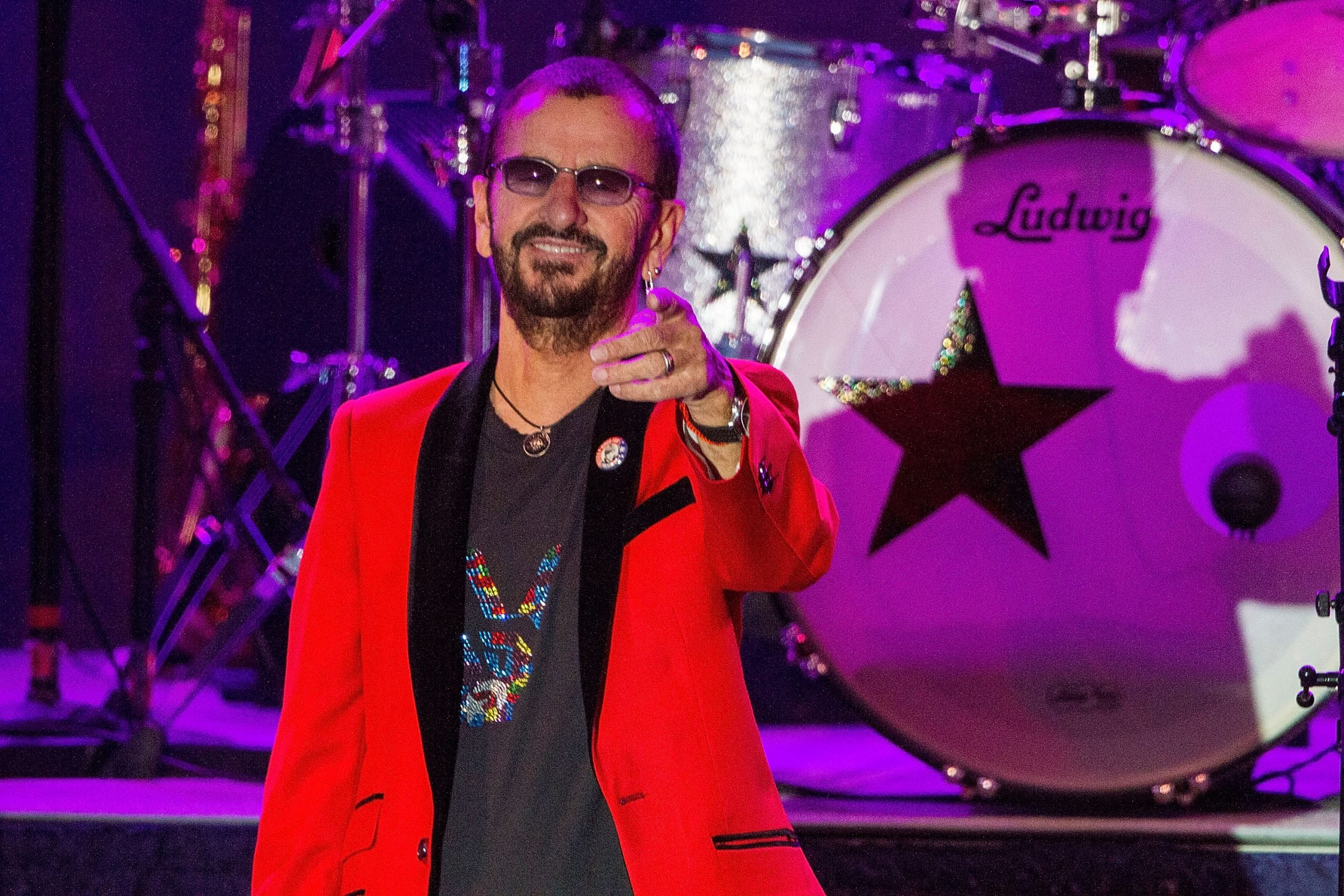 Ringo Starr wears a red jacket and T-shirt as he smiles and points out to the crowd from on stage in 2016.