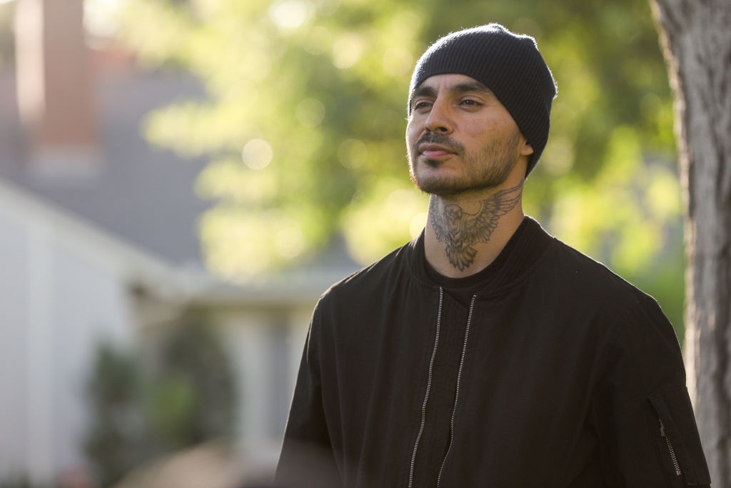 Manny Montana as Rio stands with a menacing look on his face. He's wearing a beanie and a dark jacket but his eagle tattoo is visible on his neck.