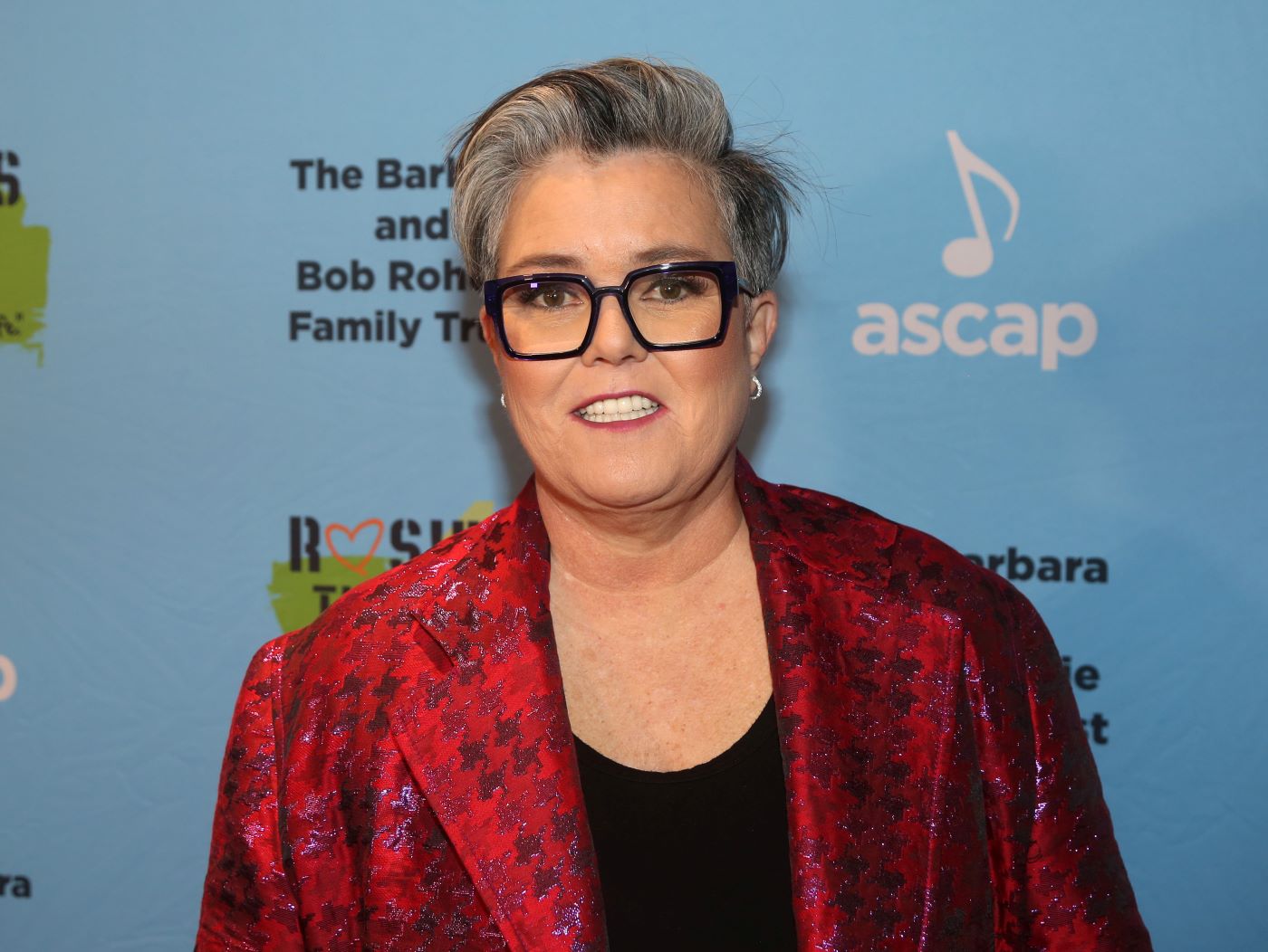 Rosie O'Donnell standing wearing a red and blank flannel suit-style jacket with a black t-shirt standing in-front of a blue background with writing in white back and green on it.