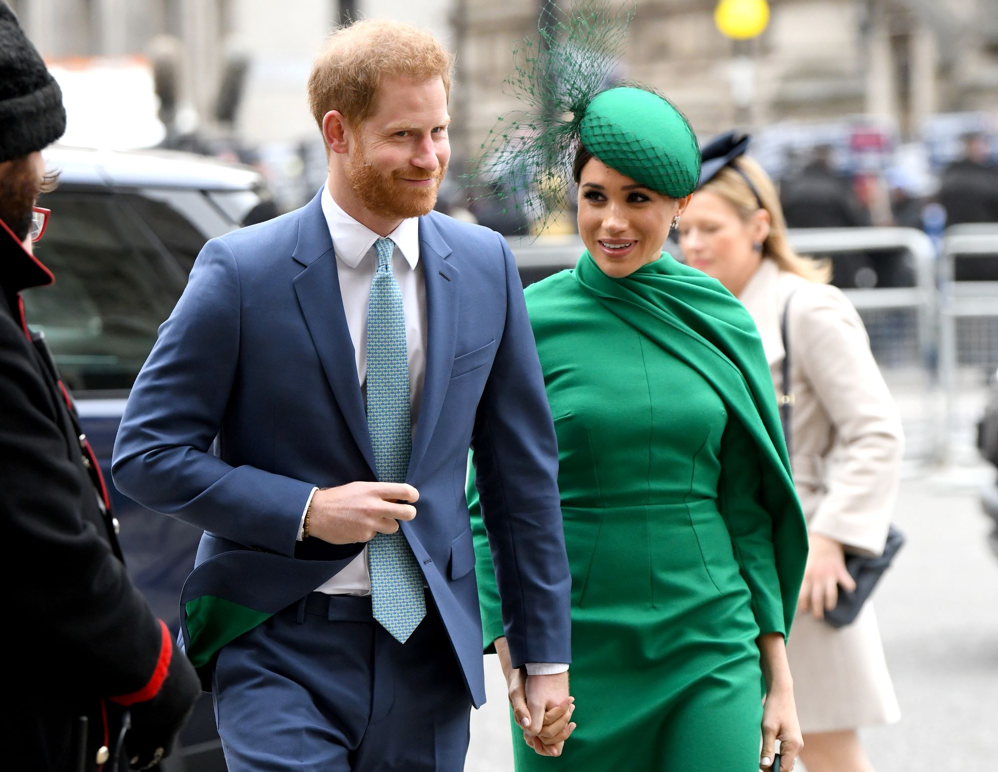 Prince Harry wears a blue suit, a white button-up shirt, a light blue tie with a darker blue pattern. Meghan Markle walks next to him, holding his hand a solid green dress and green hat. The background is a city scene with blurred cars and people walking behind.