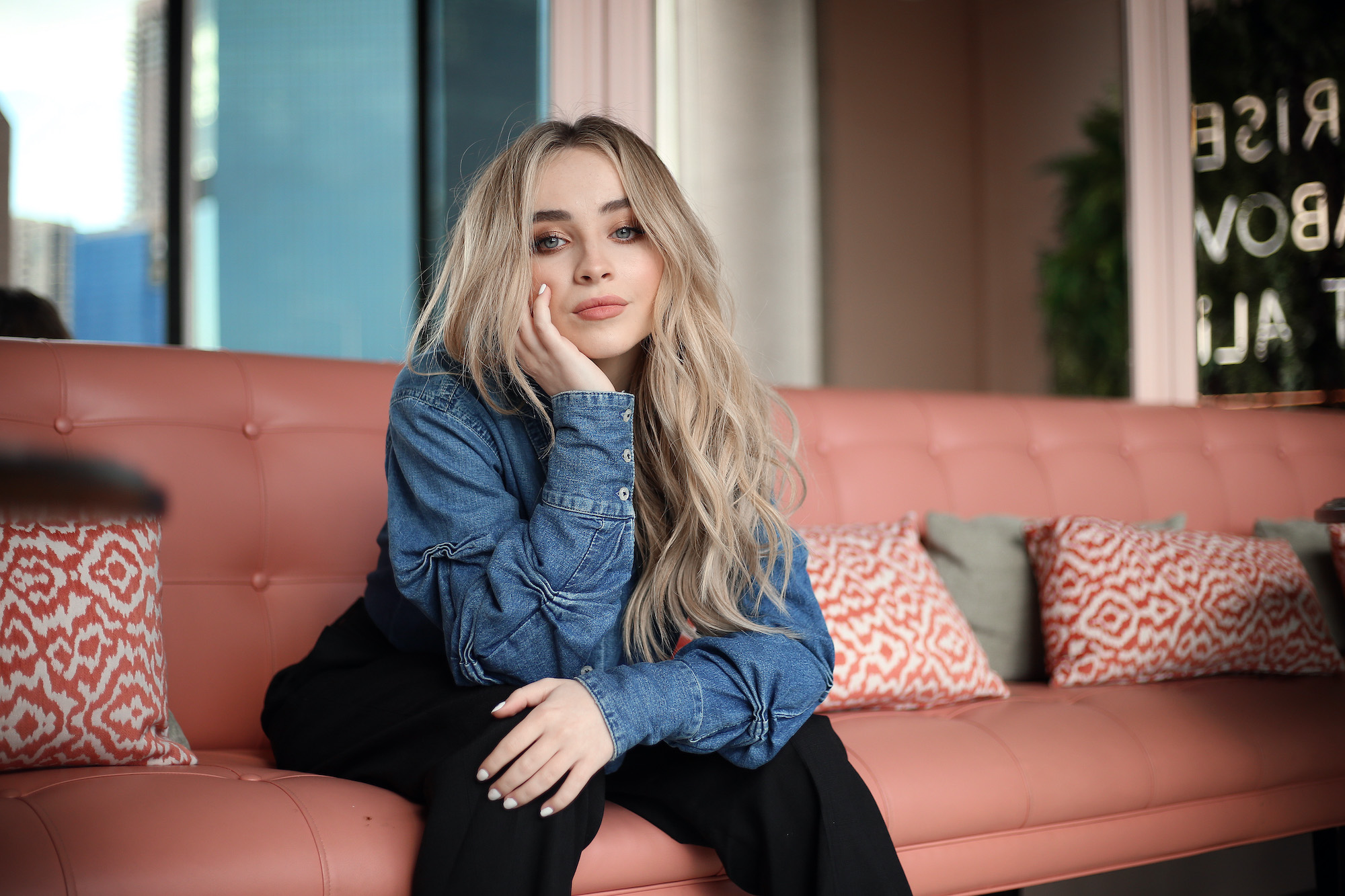 Sabrina Carpenter smiling slightly, sitting on a pink couch