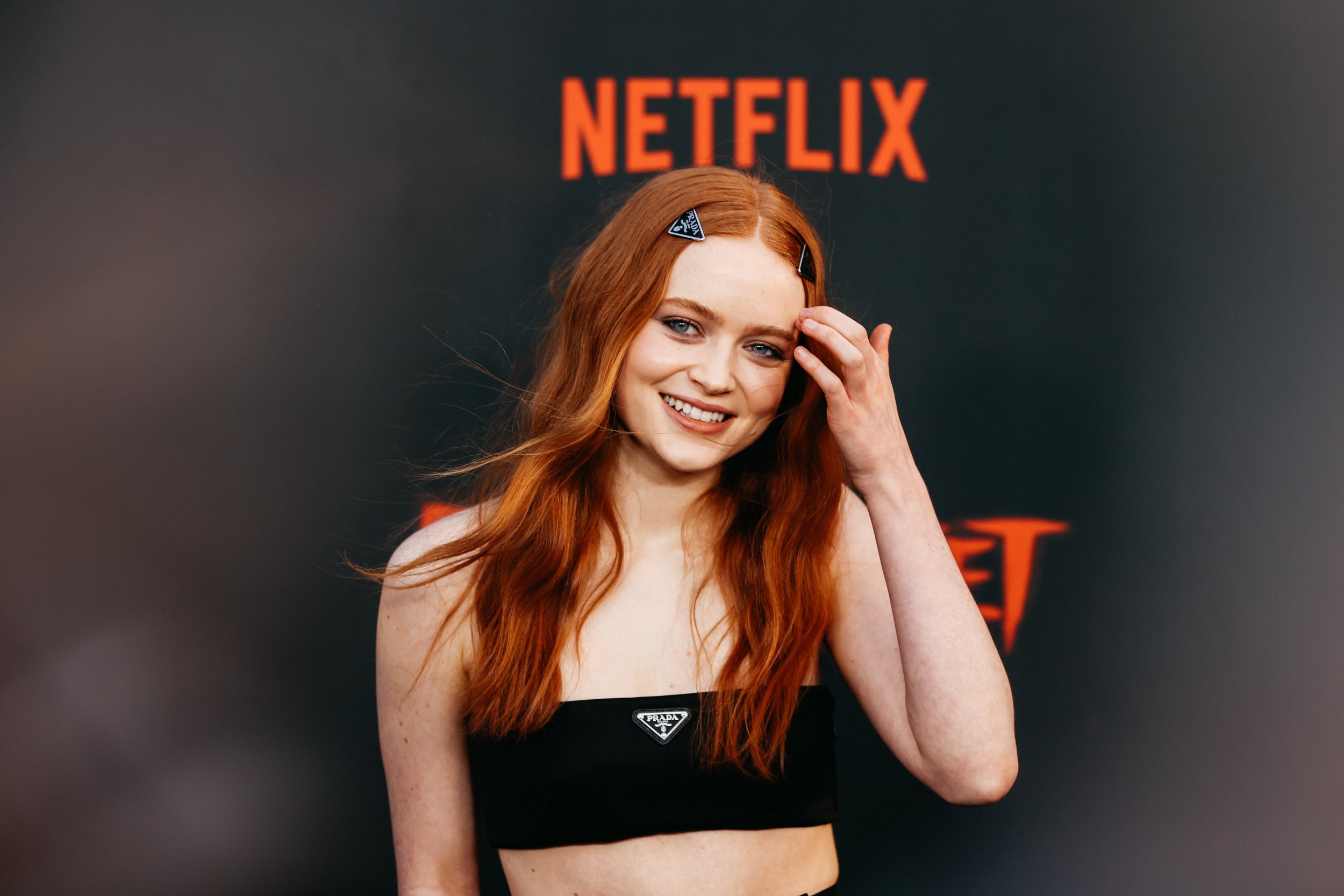 'Fear Street' star Sadie Sink smiling at the camera and touching her hair in front of a Netflix wall