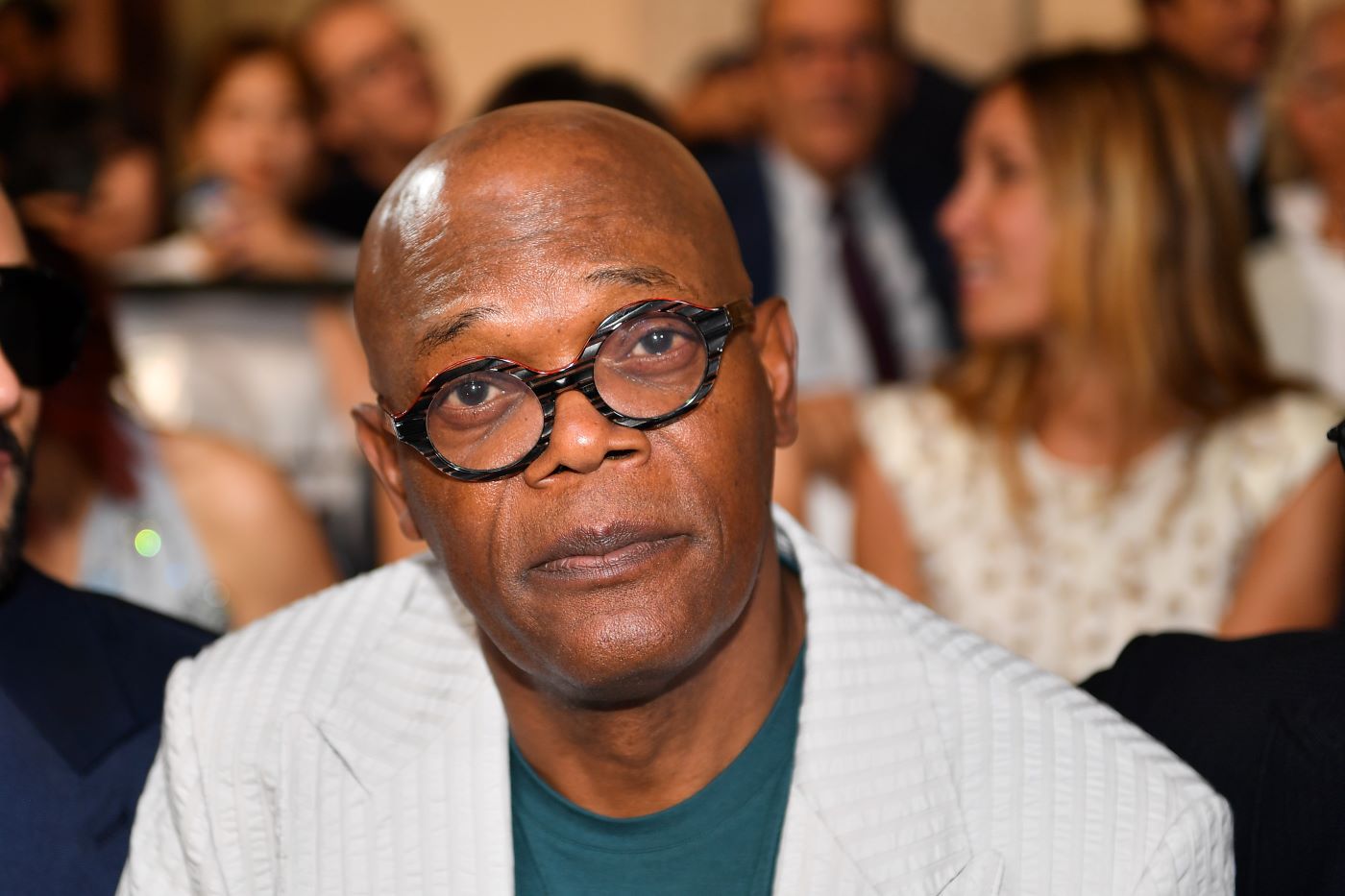 Samuel L. Jackson wearing a textured cream suit jacket with a green undershirt and black rimmed glasses in front of a background of several blurred images of people.