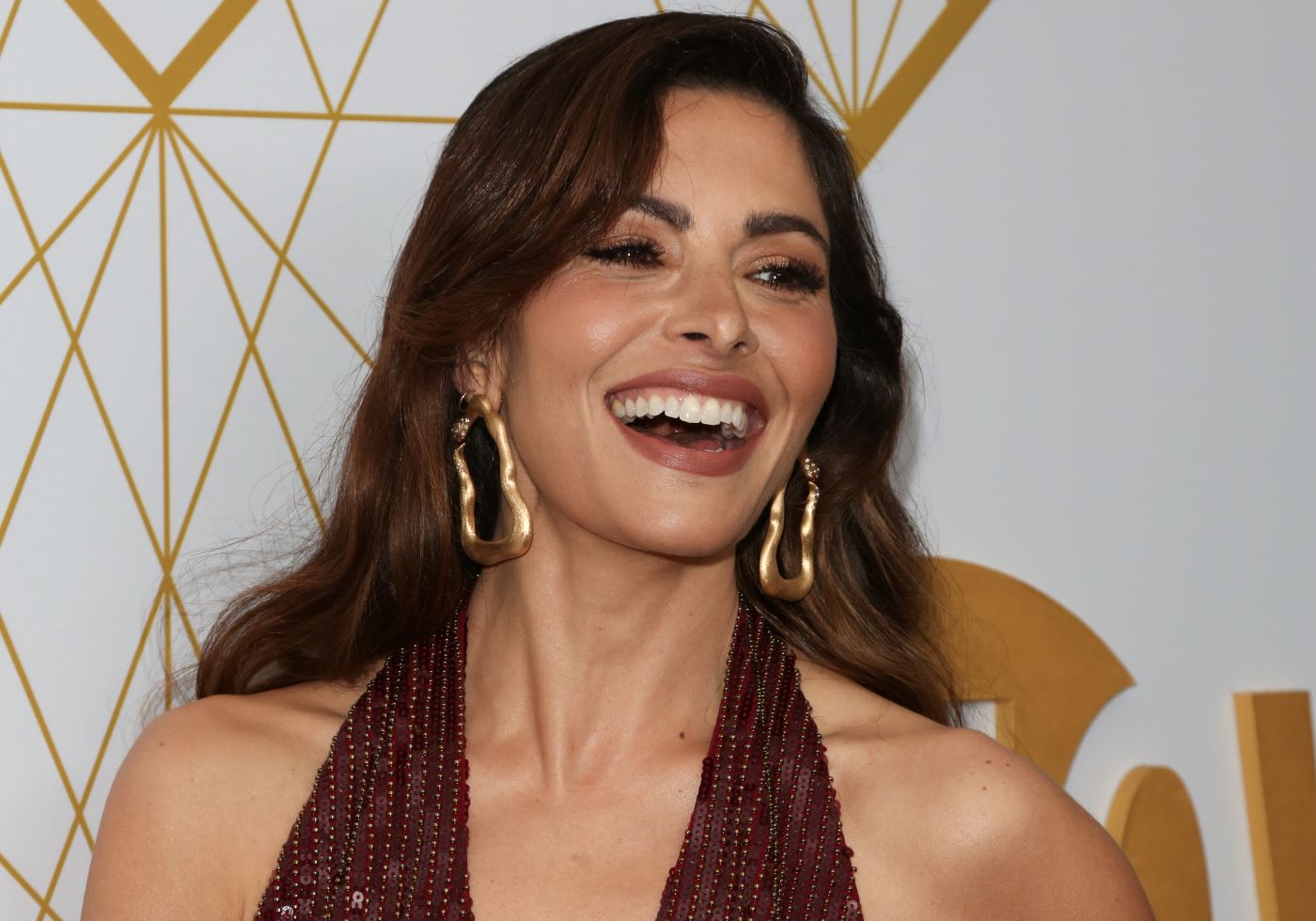 Sarah Shahi in front of a white and gold backdrop wearing a brown halter top and large gold earrings while smiling.