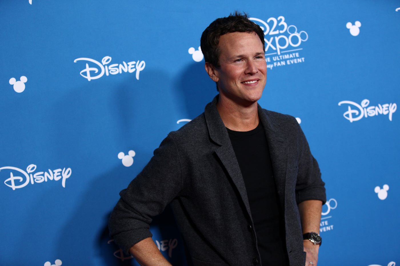 Scott Weinger, voice actor of the 1992 'Aladdin,' is standing in front of a blue backdrop with 'Disney' written in random places dressed in a black undershirt with a quarter-sleeve black suit jacket.