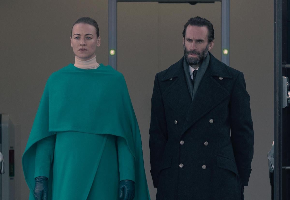 Yvonne Strahovski as Serena Joy Waterford and Joseph Fiennes as Fred Waterford in 'The Handmaid's Tale' Season 4. Strahovski wears a teal dress and coat. Fiennes wears a black wool coat and suit. They're walking out of a building with metal detectors behind them.