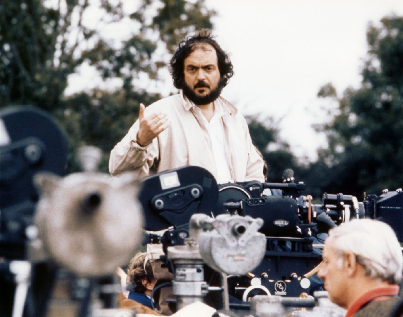 Stanley Kubrick stands behind a sea of cameras wearing a white long sleeve button up shirt with trees in the background.