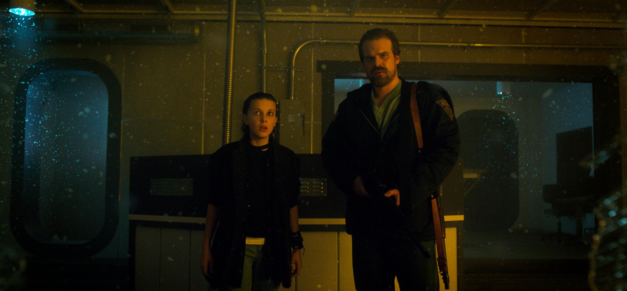 Millie Bobby Brown and David Harbour as Eleven and Hopper in Stranger Things. They're standing next to one another and Eleven is dressed in black clothes with her hair slicked back