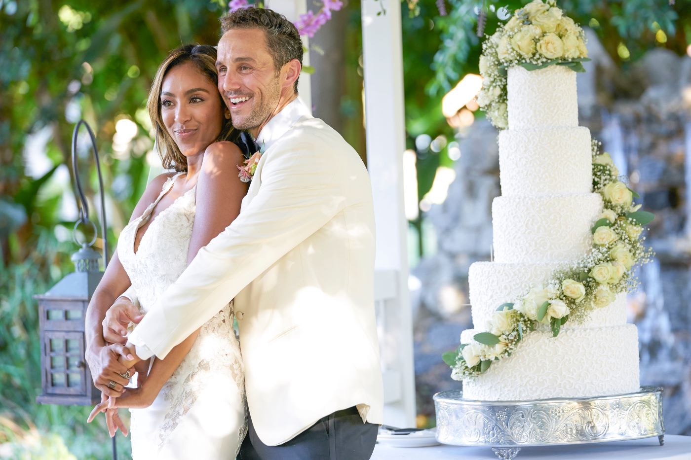 Tayshia and Zac are dressed in wedding attire, Tayshia in a white dress and Zac in a white suit, standing in front of a white wedding cake. Which 'Bachelorette' couples are still together?