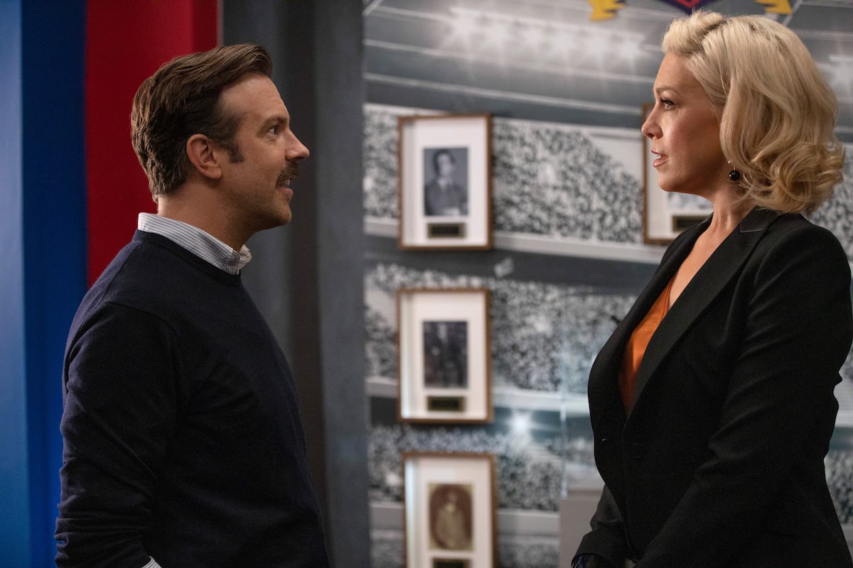 Jason Sudeikis and Hannah Waddingham in 'Ted Lasso' Season 1. Jason stands on the left in a navy blue sweater with a collared shirt underneath. Hannah stands on the right in a black suit and orange top. Their profiles face the camera. A floor-to-ceiling black-and-white photo of a crowded soccer stadium is behind them.