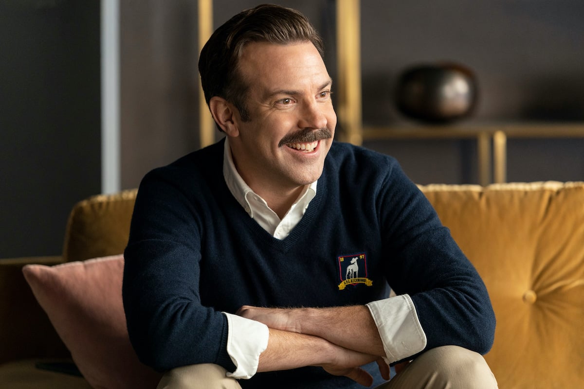 Jason Sudeikis in 'Ted Lasso' Season 2. He sits on a yellow couch with his arms crossed on his knees as he smiles. He wears a navy blue sweater with a white collared shirt underneath. Gold shelves are behind him.