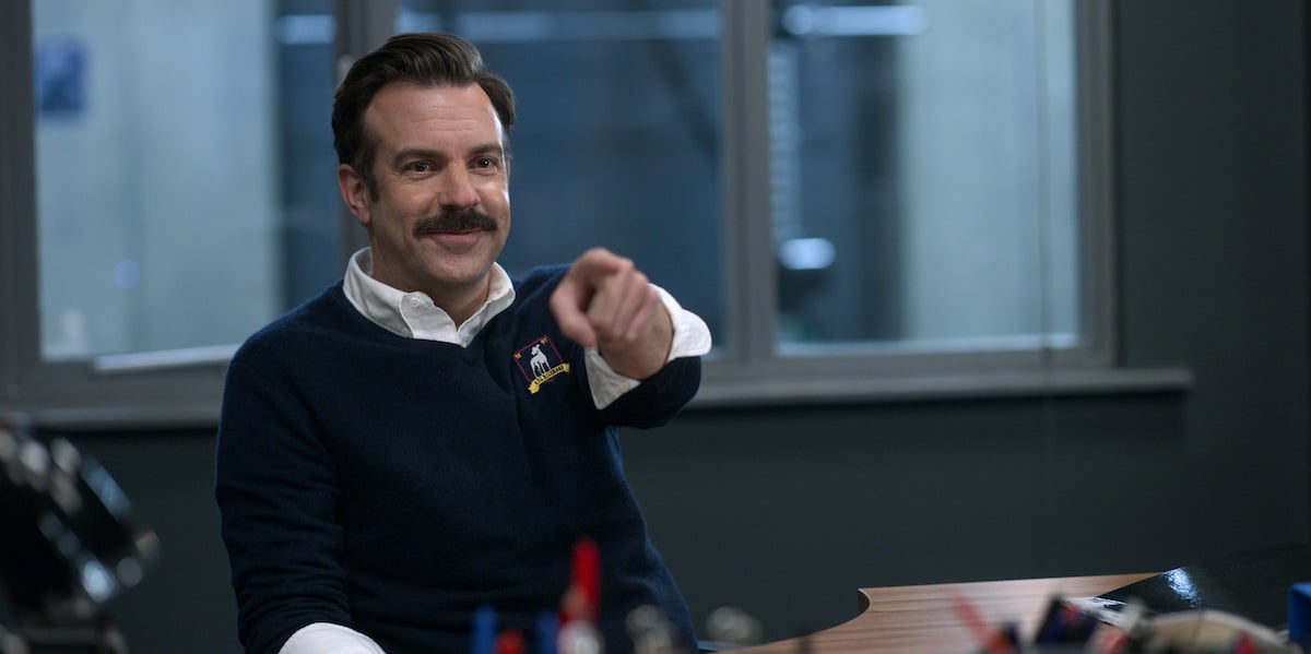 Jason Sudeikis in 'Ted Lasso' Season 2. He sits at a desk with a smirk on his face as he points to someone off camera. He sits in an office in a locker room. There's a window behind him showing the locker room.