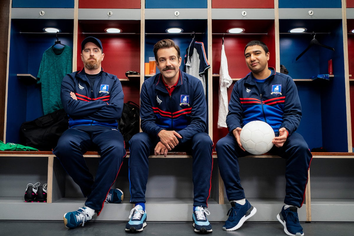 (L-R) Brendan Hunt, Jason Sudeikis, and Nick Mohammed ​in 'Ted Lasso' Season 2. The actors sit in a locker room in front of alternating blue and red cubbies. They wear blue and red athletic tracksuits and smile directly at the camera.