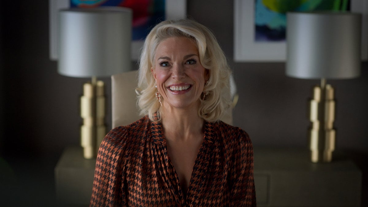 Hannah Waddingham ​in 'Ted Lasso' Season 2. She smiles in an orange and black houndstooth blouse in an office. Gold lamps and colorful paintings are behind her.