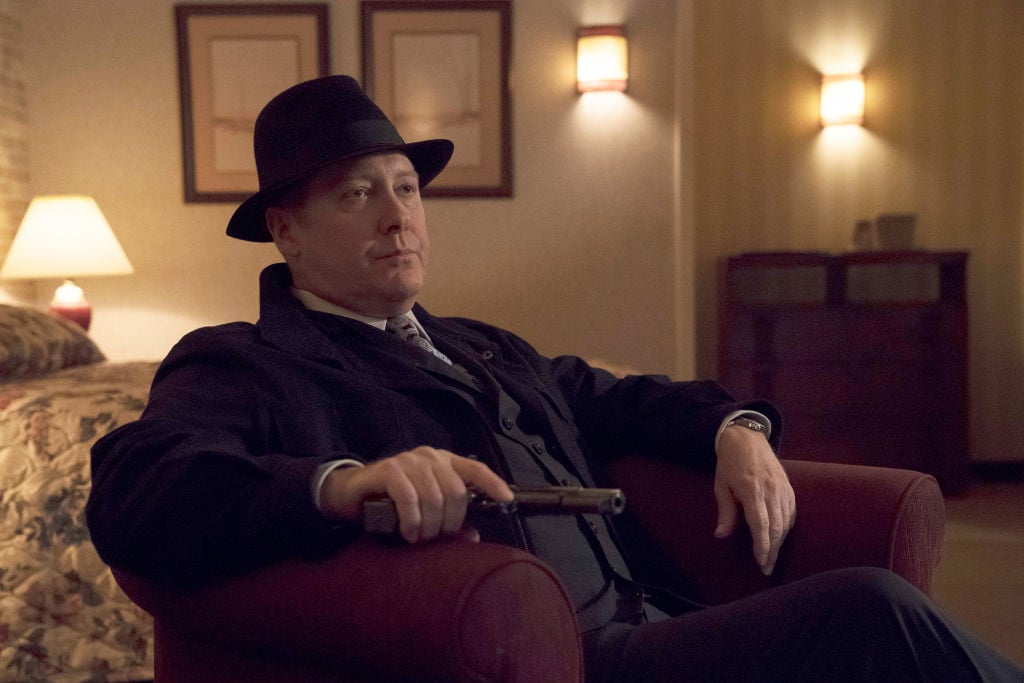James Spader as Raymond 'Red' Reddington sits in a chair, holding his gun.