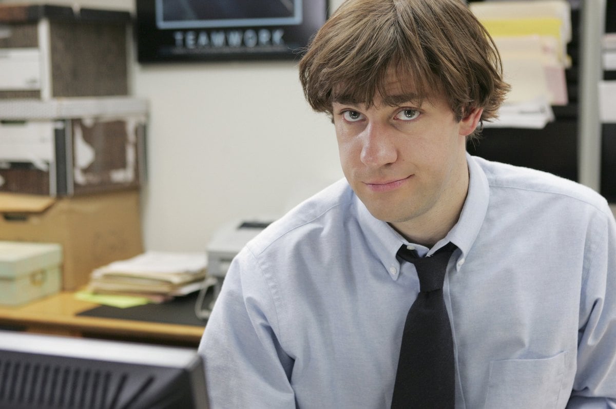 John Krasinski as Jim Halpert wears a blue shirt and a black tie as he sits in the office.
