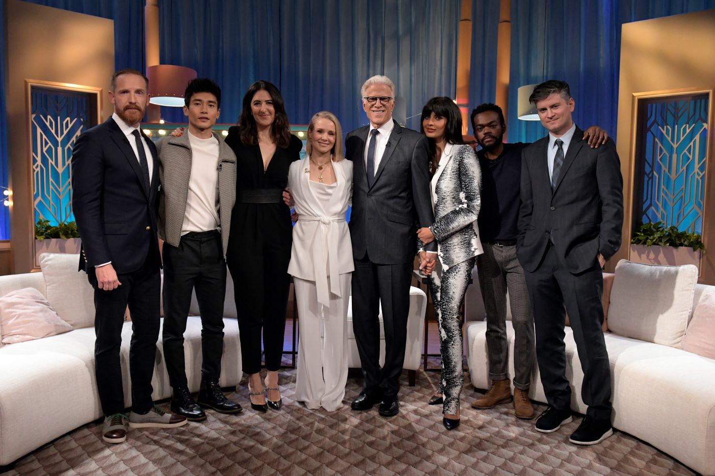 Several People from the cast of 'The Good Place' stand in a line dressed in professional attire in front of a round white couch with blue curtains and gold props in the background.