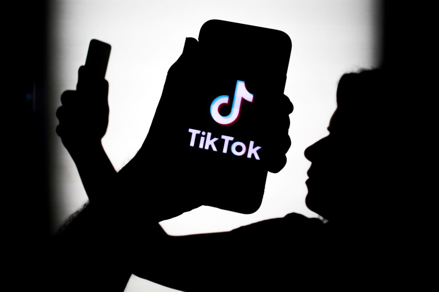 Shadows of a person holding a phone out in front of their face with their arm crossed with a closeup picture of a shadow of a hand holding a phone with the Tik Tok logo on it all against a white background.