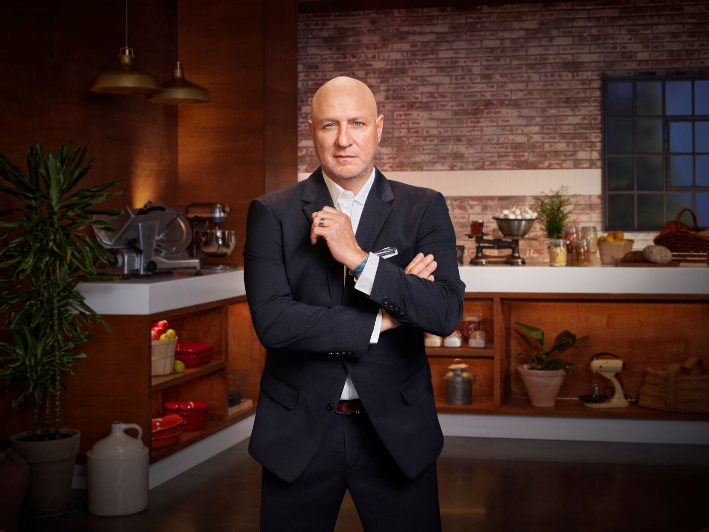 Tom Colicchio from Top Chef stands in front of the set surrounded by cookware including mixers, scales, and deli slicers in front of a half light brown brick wall and a wood wall. He is wearing a black suit with a white button-up shirt.