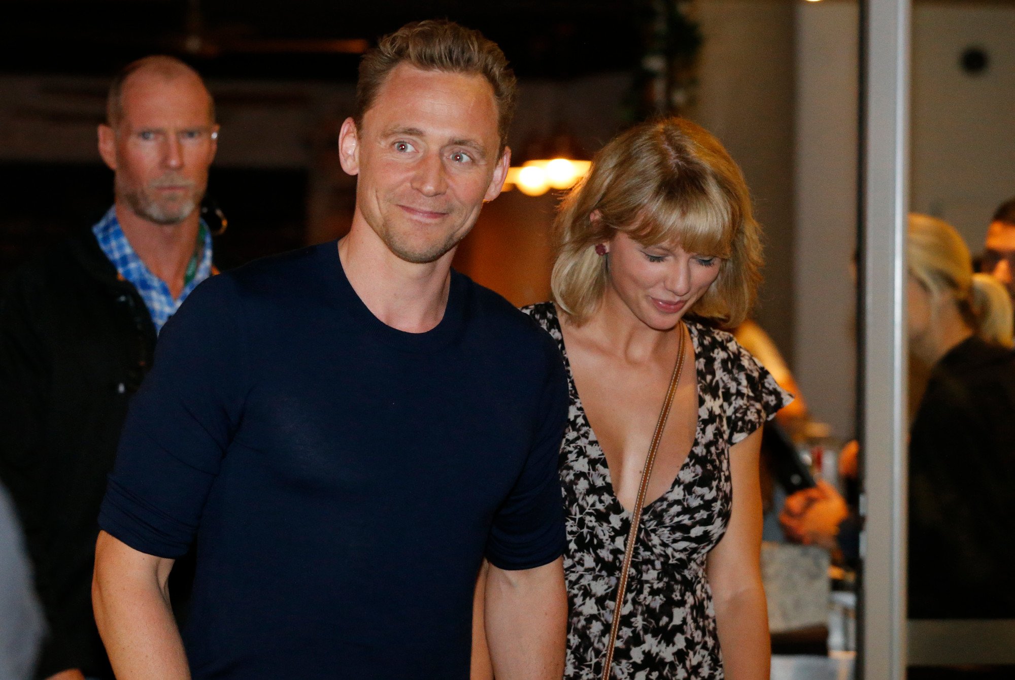Tom Hiddleston wearing a black shirt and looking surprised. Taylor Swift walks next to him with her head down.